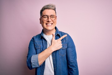 Young handsome modern man wearing glasses and denim jacket over pink isolated background cheerful with a smile of face pointing with hand and finger up to the side with happy and natural expression