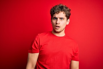 Young blond handsome man with curly hair wearing casual t-shirt over red background In shock face, looking skeptical and sarcastic, surprised with open mouth