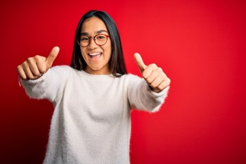 Young beautiful asian woman wearing casual sweater and glasses over red background approving doing positive gesture with hand, thumbs up smiling and happy for success. Winner gesture.