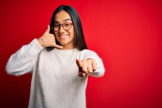 Young beautiful asian woman wearing casual sweater and glasses over red background smiling doing talking on the telephone gesture and pointing to you. Call me.