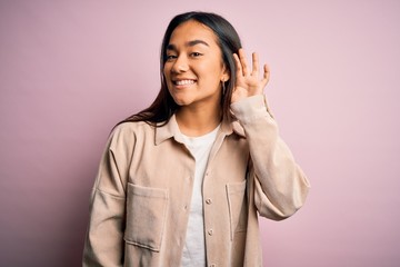 Young beautiful asian woman wearing casual shirt standing over pink background smiling with hand over ear listening an hearing to rumor or gossip. Deafness concept.