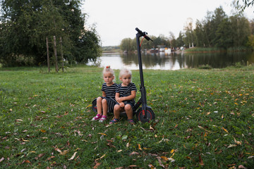 Twin blonde little girls sit on fallen electric scooter in green park