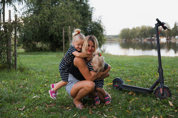Twin blonde little girls sitting on electric scooter in park with their mother