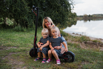 Twin blonde little girls sitting on electric scooter in park with their mother