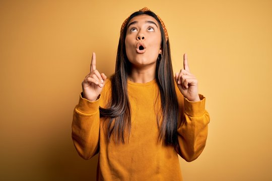 Young beautiful asian woman wearing casual sweater and diadem over yellow background amazed and surprised looking up and pointing with fingers and raised arms.