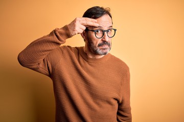 Middle age hoary man wearing brown sweater and glasses over isolated yellow background pointing unhappy to pimple on forehead, ugly infection of blackhead. Acne and skin problem