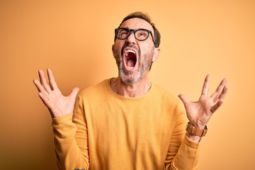 Middle age hoary man wearing casual sweater and glasses over isolated yellow background crazy and mad shouting and yelling with aggressive expression and arms raised. Frustration concept.