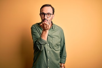 Middle age hoary man wearing casual green shirt and glasses over isolated yellow background looking at the camera blowing a kiss with hand on air being lovely and sexy. Love expression.