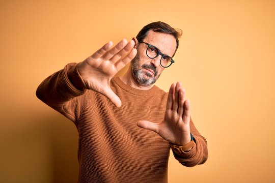 Middle age hoary man wearing brown sweater and glasses over isolated yellow background doing frame using hands palms and fingers, camera perspective