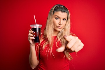 Young beautiful blonde woman drinking cola fizzy beverage using straw over red background pointing with finger to the camera and to you, hand sign, positive and confident gesture from the front