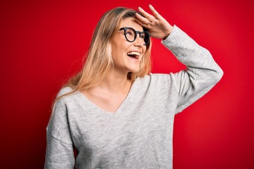 Young beautiful blonde woman wearing sweater and glasses over isolated red background very happy and smiling looking far away with hand over head. Searching concept.