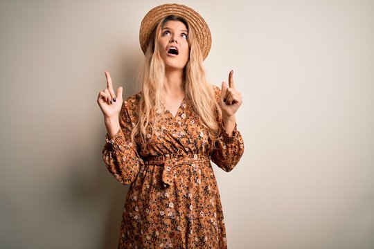 Young beautiful blonde woman wearing summer dress and hat over isolated white background amazed and surprised looking up and pointing with fingers and raised arms.