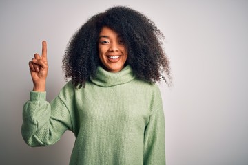 Young beautiful african american woman with afro hair wearing green winter sweater showing and pointing up with finger number one while smiling confident and happy.