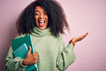 Young african american student woman with afro hair reading books over pink background very happy...