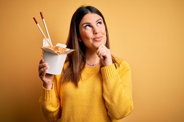 Young beautiful woman eating asian ramen noodles using chopsticks over yellow background serious face thinking about question, very confused idea