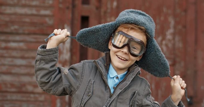 Portrait shot of Caucasian cute small boy in hat and glasses smiling to camera and having fun in costume aviator outdoor. Close up of funny little red-haired kid laughing and playing in pilot.