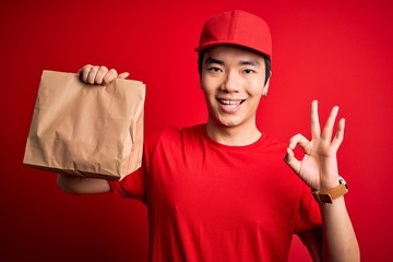 Young handsome chinese delivery man holding takeaway paper bag with food doing ok sign with fingers, excellent symbol