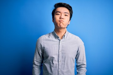 Fototapeta premium Young handsome chinese man wearing casual shirt standing over isolated blue background puffing cheeks with funny face. Mouth inflated with air, crazy expression.