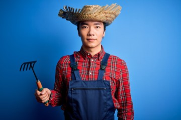 Young handsome chinese farmer man wearing apron and straw hat holding rake tool with a confident expression on smart face thinking serious