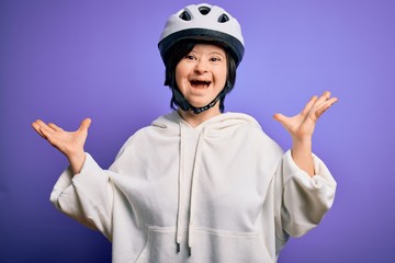 Young down syndrome cyclist woman wearing security bike helmet over purple background celebrating crazy and amazed for success with arms raised and open eyes screaming excited. Winner concept