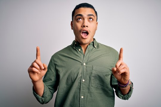Young brazilian man wearing casual shirt standing over isolated white background amazed and surprised looking up and pointing with fingers and raised arms.
