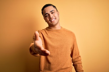 Young brazilian man wearing casual sweater standing over isolated yellow background smiling cheerful offering palm hand giving assistance and acceptance.