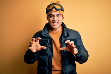 Young brazilian skier man wearing snow sportswear and ski goggles over yellow background smiling funny doing claw gesture as cat, aggressive and sexy expression