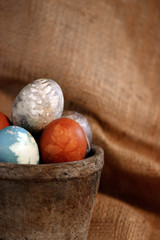 A collection of natural-dyed eggs with botanical leaf prints in a rustic gray clay pot against a draped burlap background, with copy space