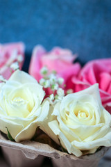 Fresh white and pink hybrid tea rose flowers and white baby's breath (Gypsophila) displayed in an egg carton against a denim blue background, with copy space