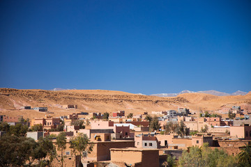 Fototapeta premium Berber village full of trees and sand lies at bottom of the desert hill with the mountains background