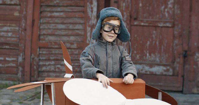 Cute funny small Caucasian boy with red hair in hat and glasses sitting outdoors in wooden toy of airplane and walking the yard. Happy child dreaming to be pilot when grow up.