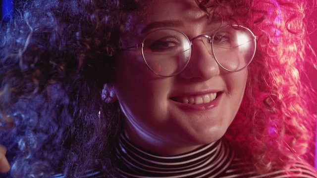 Neon girl portrait. 80s style. Shy woman in eyeglasses playing with curly hair in blue pink lights.