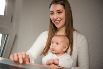 Young mother holds baby girl and works on laptop computer