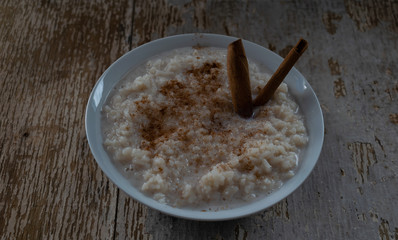 Fototapeta na wymiar postre de arroz con leche, espolvoreado con canela en polvo y dos palitos de canela, en un plato blanco, sobre tabla de madera