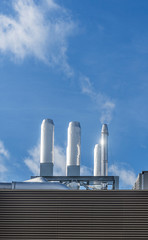 Shiny metal exhaust pipe chimneys on a building rooftop against a clear blue sky, emitting white particles.