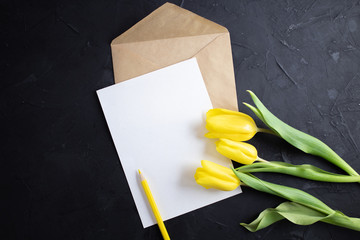 Yellow tulips on the edge of white paper for text on a dark background. A blank sheet of paper with an envelope. Copy space.