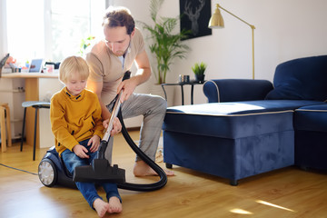 Fototapeta na wymiar Young handsome father teaches his son how to use a vacuum cleaner. Loving dad and his preschooler boy doing housework together at home.