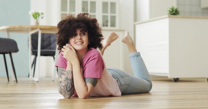 Happy pretty girl with curly hairs at home lying on floor looking at camera and smiling. Brunette woman is resting. Charming caucasian girl in apartment.