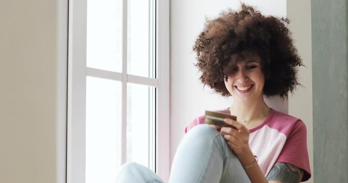 Happy woman with curly hairs is making online payment holding bank card using smartphone at home. Finance, online shopping in internet concept. Doing online purchase sitting near window.