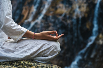 Close hand of a woman in white clothes meditating on a rock by a waterfall