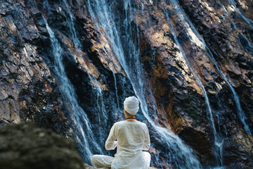 Young meditating woman in a white clothes and turban sitting on the rock by the waterfall in the forest