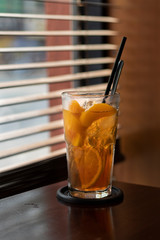 peach iced tea in a glass with slices of peach, wooden table and window blinds in the background