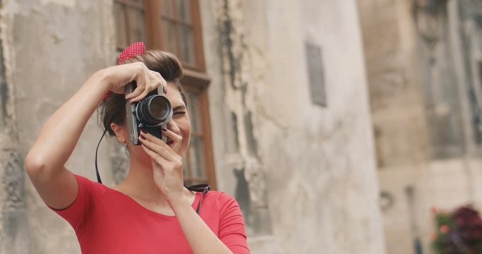 Portrait of vintage good looking Caucasian woman smiling while taking photos on retro photocamera at daytime on the street. Charming young trendy girl in red blouse with camera outdoors Trend concept