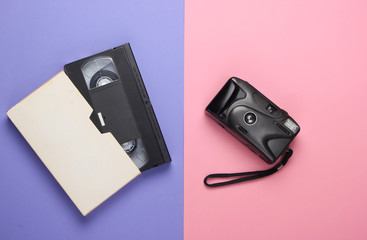 Video cassette and retro camera on pink-purple pastel background. Top view