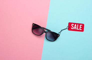 Fashionable sunglasses with red sale tag on pink blue pastel background. Top view. Discount....