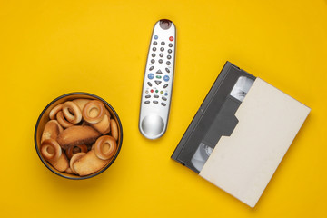 TV remote control and bowl of crackers, video cassette on yellow background. Watching the TV. The...