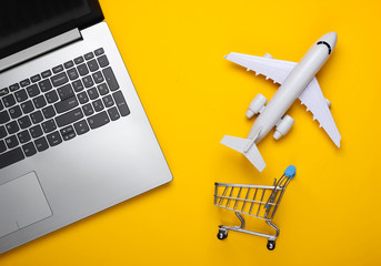 Flat lay composition. Air delivery. Online shopping. Laptop, figurine of shopping trolley, airplane on yellow background. Top view