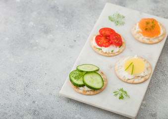 Various round healthy crackers with salmon and cheese, tomato and cucumber on marble board on light kitchen table background. Space for text