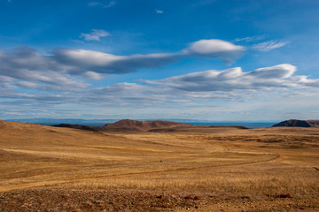 Steppe with hills, clouds in the sky and mountains in the distance. Country road. Brown and yellow grass. Blue sky with fluffy clouds.