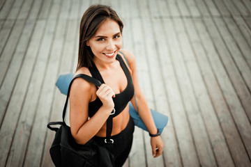 Slender cheerful fit woman in sport clothes with gym bag and mat on her shoulders posing after training outdoors